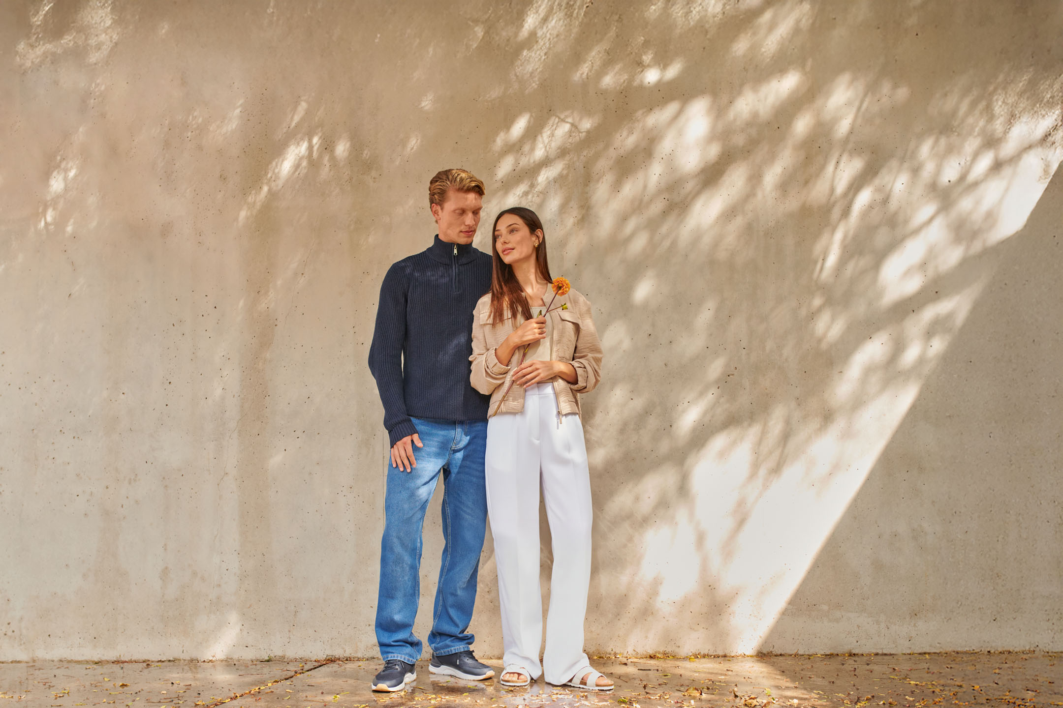 Man in donkerblauwe trui en jeans, vrouw in beige jasje en witte broek met bloem.