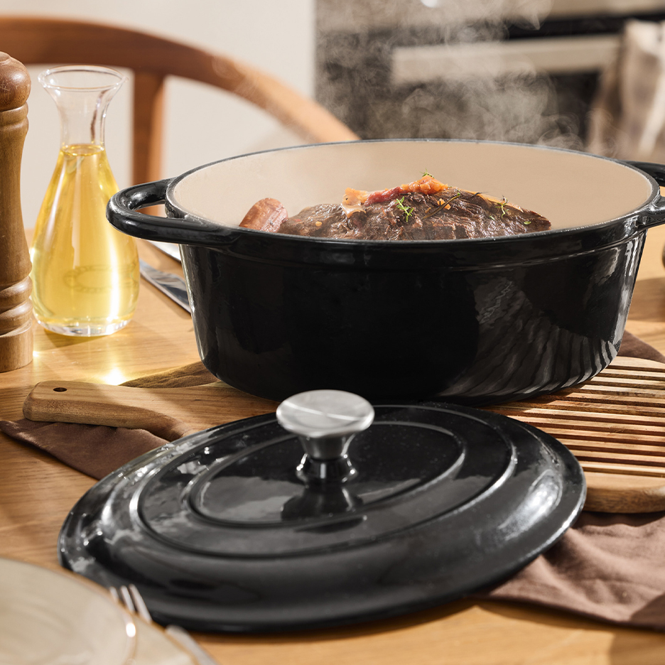Black oval cast iron pot with steaming meat dish on a wooden table.