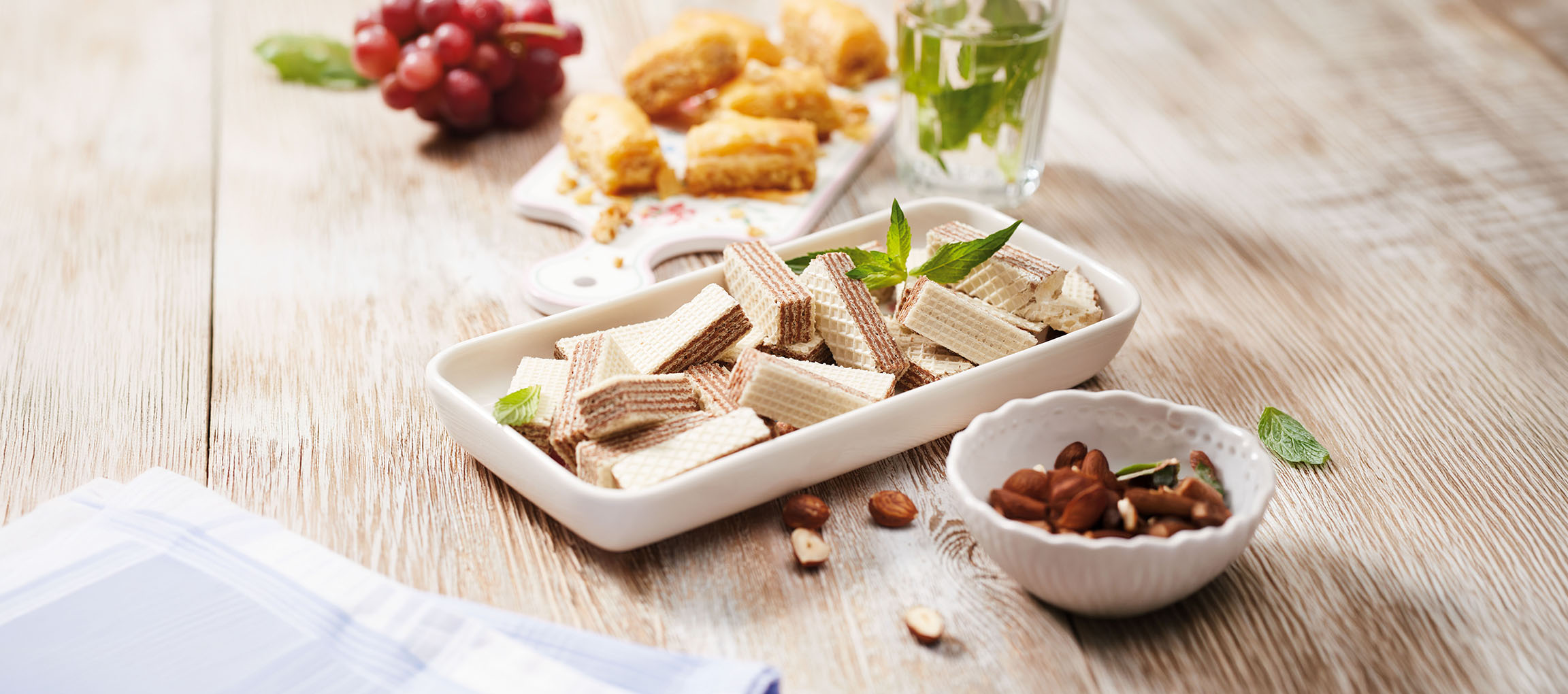 Wafer biscuits, baklava, grapes, and mint water on a wooden table.
