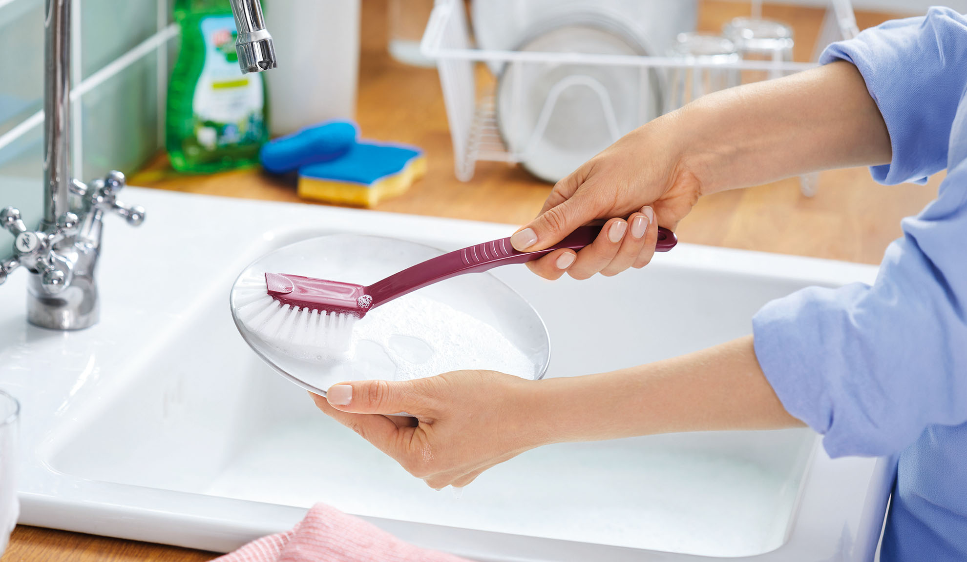 Woman washing dishes in a sink with a dish brush and plate, dish rack visible.