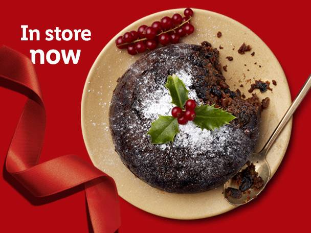 A Christmas pudding with holly and red berries on a plate, with a red ribbon and text 'In store now'.