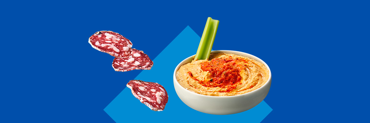 Salami slices and a bowl of hummus with a celery stick on a blue background.