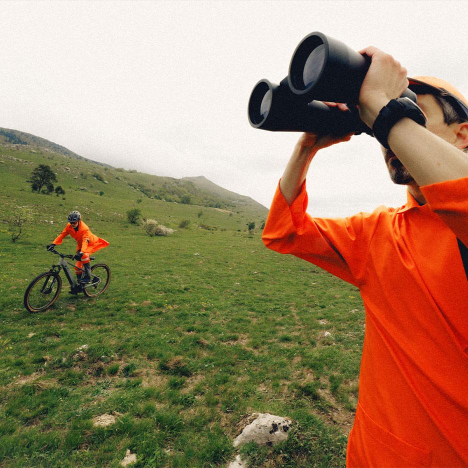 Man with binoculars and cyclist in orange sportswear in a mountain landscape.