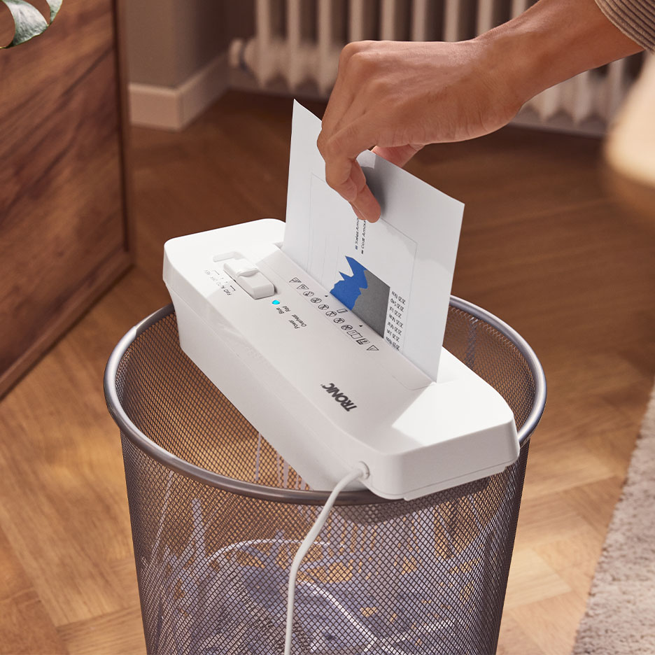Hand feeding paper into a TRONIC paper shredder placed on top of a waste bin.