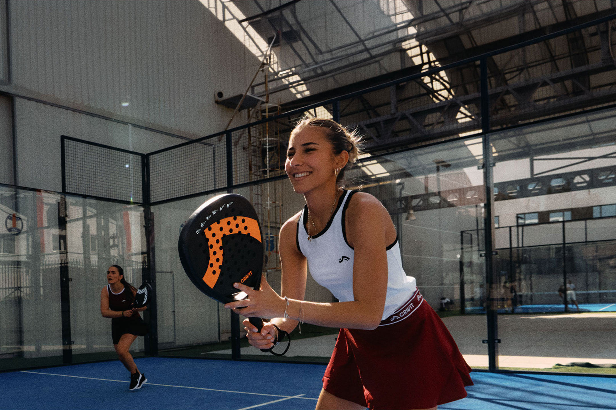 aiLabeling.aiLabelForImageAltText: Two women playing padel tennis, one smiling and holding a racket, on a blue court.