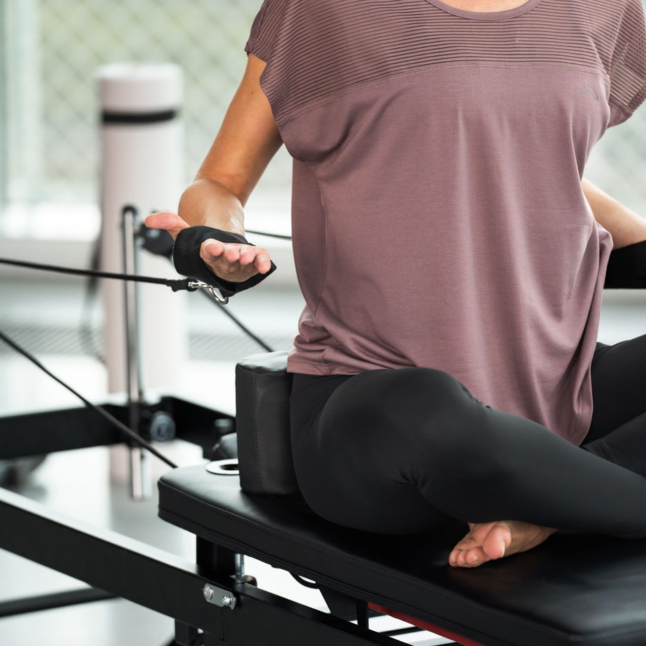 Woman exercising Pilates on a reformer, wearing black leggings and a purple top.