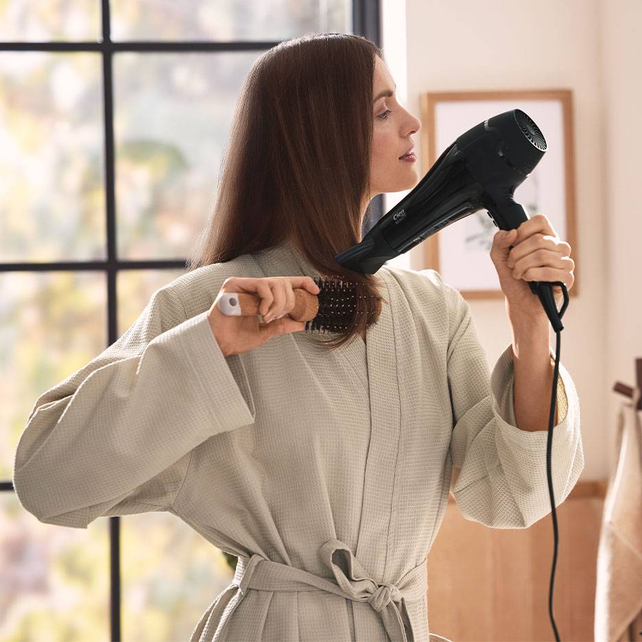 Woman drying her hair with a hairdryer and round brush, wearing a bathrobe.