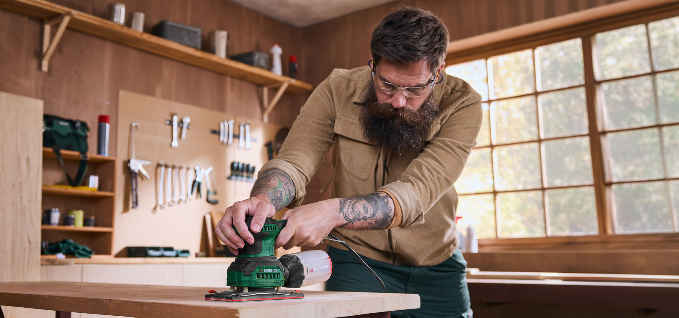Man sanding a wooden board with an electric sander in a workshop.
