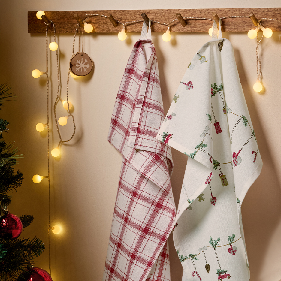 Two festive Christmas kitchen towels, one plaid and one with holiday motifs, hanging on a wooden hook rack with string lights.