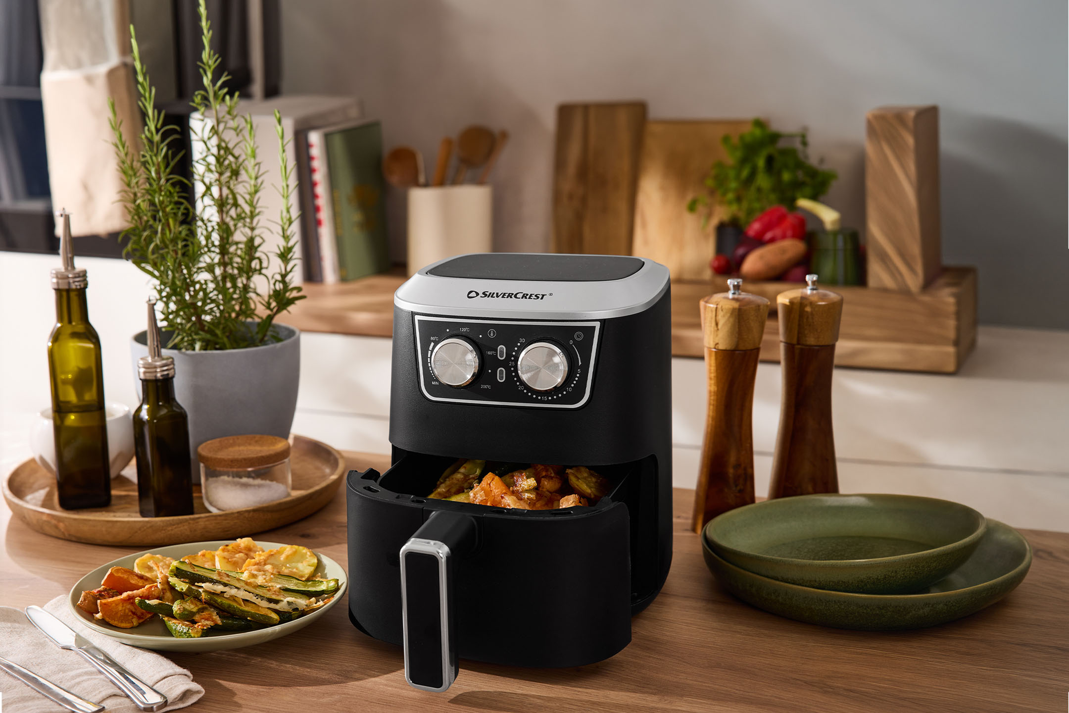 Black air fryer with vegetables, spices, and dishes on a wooden countertop.