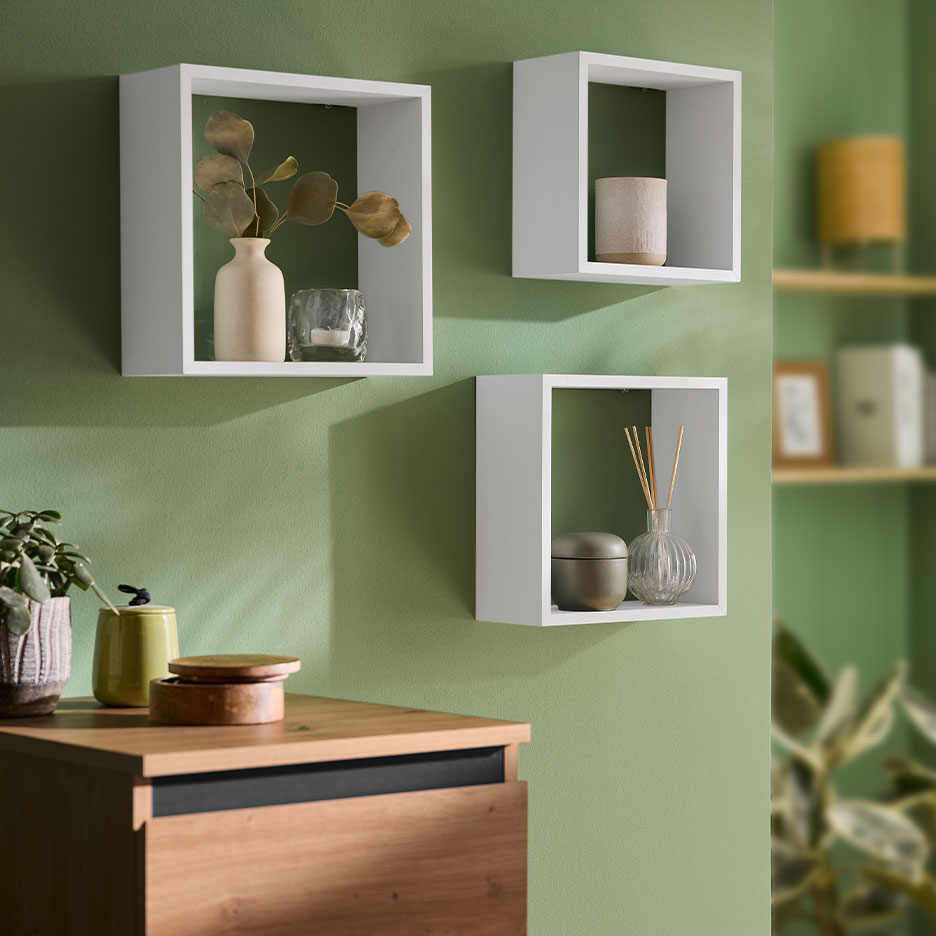 White wall shelves with decorations on a green wall, with a wooden chest of drawers underneath.