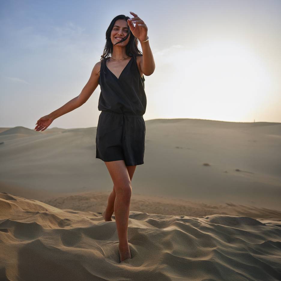 Woman in black jumpsuit barefoot standing on sand dunes at sunset.