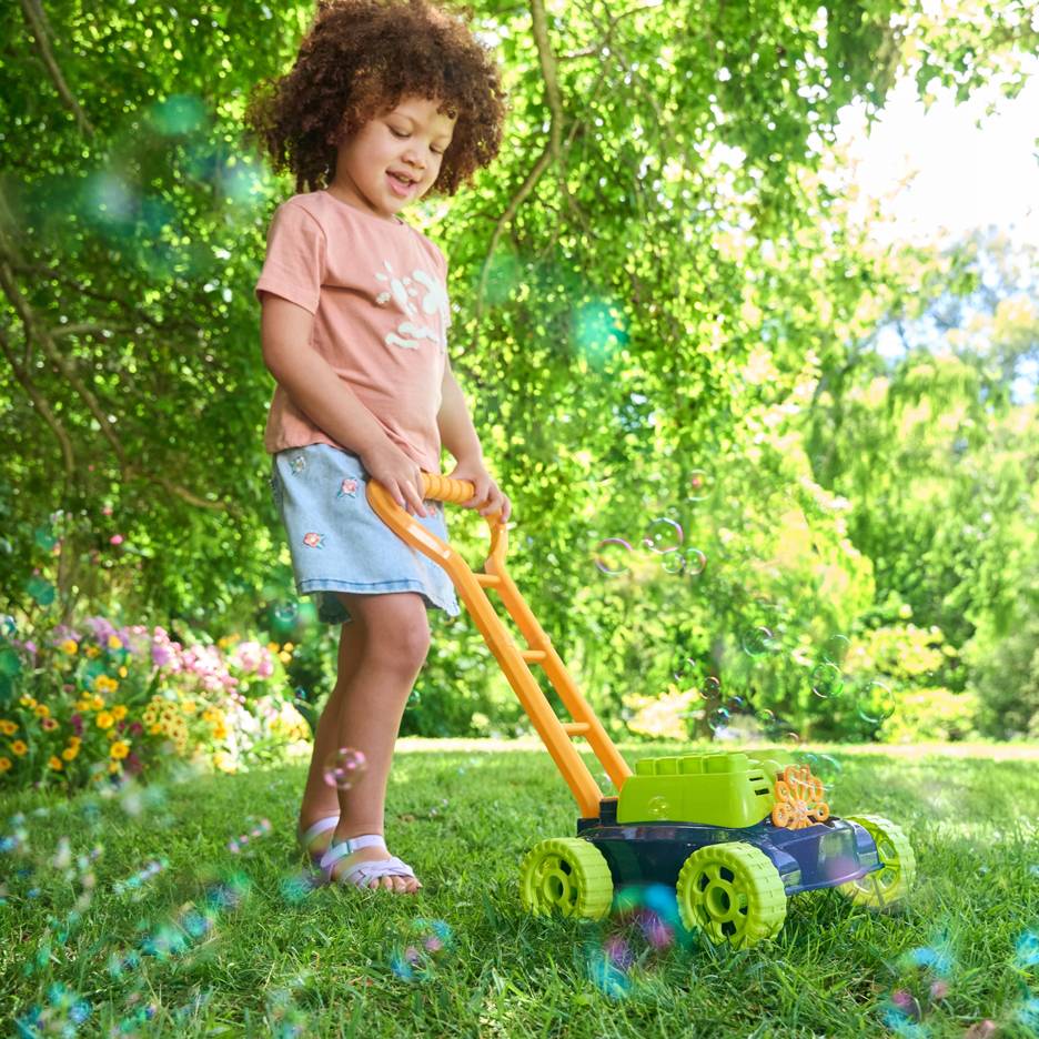 A child with curly hair pushes a toy bubble lawnmower on green grass.